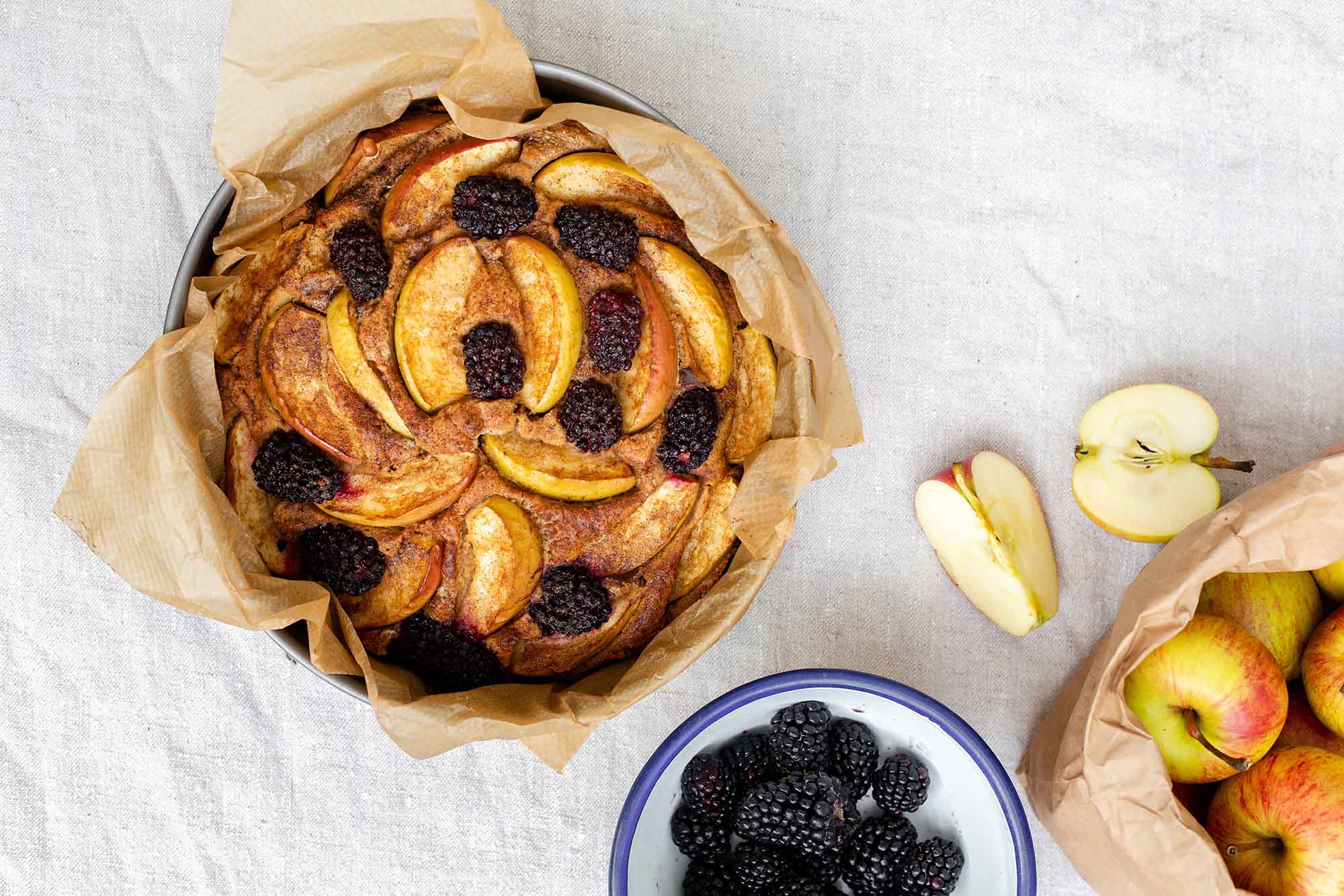 An apple and blackberry cake in a tin next to a bowl of blackberries and apples