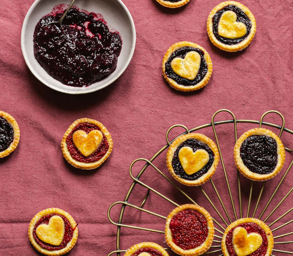 Berry chia jam tarts with hearts on a pink tablecloth next to some berries in a bowl